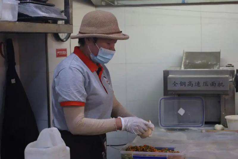 什么是&ldquo;义乌精神&rdquo;？65岁的邵宝玲有自己的答案。  上世纪80年代，她是义乌第一批&ldquo;万元户&rdquo;，后来创办箱包企业，年销售额数亿。2014年前后，因为决策失误替人担保，她的资金链断裂，负债累累。走投无路时，母亲一句&ldquo;那就去做包子&rdquo;让她重新振作。如今，包子铺开到第三家。她说，&ldquo;跌倒了不可怕，站起来，都有机会。&rdquo;  时光里｜人生不止一个回合 凌晨三点，义乌街头一片墨色。 邵宝玲的一天从这时开始。一辆老旧的货车驶出梧桐树影，她坐上副驾驶，系好安全带，目的地是十二公里外的蔬菜批发市场。 六十五岁。曾经的&ldquo;箱包女王&rdquo;，现在的包子铺老板&ldquo;宝玲姐&rdquo;。四年前这个点，她躺在出租屋里数天花板上的裂纹；现在，她正低头认真挑选包包子的南瓜。她说，命运总是公平的。曾经那么爱睡懒觉的人，如今凌晨三点起床；曾经风光无两的老板，最后去卖了包子。  邵宝玲在菜市场买菜 手还是那双手，茧子换了位置。 四十年前，这双手数过一摞摞的现金&mdash;&mdash;1986年，邵宝玲和丈夫一起创办装潢公司，成为义乌第一批&ldquo;万元户&rdquo;。一万块钱，她翻来覆去数了三遍，手都在抖。 二十多年前，这双手摸过一层层皮革&mdash;&mdash;2002年，她创办箱包公司，2600多个箱包品种，年销售额数亿元。订单从全世界涌来，流水线日夜不停。 她曾想赚到十万就退休，结果十万很快赚到；又想赚到一百万就不干了，一百万也很快来了。后来她不再说这话，只是一直干下去。  邵宝玲在她引以为傲的箱包博物馆中讲解 邵宝玲参加行业活动 2012年，浙江省两会。邵宝玲坐在大会堂里，穿着得体的套装，胸前别着红色的代表证。她说&ldquo;选上了就要做表率，一举一动都要做好。&rdquo;她是这么说的，也是这么做的：资助贫困学生，给员工盖宿舍，借钱给同行周转。她信奉义乌人的生意经&mdash;&mdash;&ldquo;出六归四&rdquo;，赚了十块钱，要拿出六块分给别人。  2012年，邵宝玲作为浙江省人大代表参加浙江省两会 &ldquo;凭手艺吃饭，不会亏到哪里去&rdquo; 但商海浮沉。2014年，因替人担保，她的资金链骤然断裂。&ldquo;看着别人出事时还在观望，等火烧到自己裤脚跟，已经晚了。&rdquo; 2018年，她一手打造的电商产业园宣告破产；2022年，成立整整二十年的巨龙箱包正式关门。所有资产都被拿来还债，&ldquo;富翁&rdquo;成了&ldquo;负翁&rdquo;。最难的时候，她把自己关在家里，窗帘拉上，不见人，不接电话。她站在出租屋的窗边，往下看了无数次，想过跳楼，一了百了。 最终没有跳。她想通了：活下来才有机会。 真正让她站起来的，还是母亲的一句话。母亲上世纪八十年代在供销社食堂掌勺，&ldquo;邵家包子&rdquo;和&ldquo;芝芝拉面&rdquo;在义乌城西远近闻名，一门手艺养活了一家人。母亲告诉她，无论什么年代，凭手艺吃饭，货真价实，都不会亏到哪里去。她决定，重新拾起母亲的衣钵，开一家包子铺。 她开始找店面。好位置的店很多，但一问租金，十几万，她一分钱都掏不出来。 转机出现在一个普通的下午。九个要好的小姐妹来看她，她做了包子招待。姐妹们连声说好吃，她鼓起勇气说想开包子铺重新创业。姐妹们当场你两万我两万，凑了十八万。 这十八万，成了六十一岁的邵宝玲从头再来的全部底气。  邵宝玲正在做包子 &ldquo;义乌人不怕生意小，只怕站不起来&rdquo; 2022年11月25日，第一家宝玲姐包子铺开业了。 有人悄悄来看热闹：当年的箱包女王，开这么个小包子铺，能成吗？她用行动给出了答案：义乌人不怕生意小，只怕站不起来。 她每天凌晨三点起床，买菜、调馅、揉面，一天干满十八个小时。为了省钱，她和员工在包子铺二楼的地上睡了四个月，铺一层海绵当床，天花板往下滴水，被子永远是潮的。 她的包子比别家的大一圈。别人卖两块五，她卖五块。这套&ldquo;定价理论&rdquo;是当年做全球箱包贸易时悟出来的，如今用在了面粉和肉馅上。她说，&ldquo;定位定价是定天下，包子是给那些跟我一样跌倒过、就想吃一口干净热乎饭的人吃。人家卖两块五我也卖两块五，那是抢别人的饭碗。我卖五块，但我做得大、料足、干净。&rdquo; 闲暇时，她架起直播设备，拍包子的制作过程。她说自己不是想当网红，就是想让别人知道，这包子干干净净的。就像她这个人，跌倒了爬起来，还是干干净净的。 直播镜头打开，有人认出了她。有一条留言她看了很久，上面写着：&ldquo;人生不只有一个回合。&rdquo; &ldquo;企业可以破产，人品不能破产&rdquo; 不是非要赢，而是输得起；不是要躺平，而是随时都有再来一次的勇气。她说，这就是义乌精神。义乌人从鸡毛换糖开始，挑着担子走街串巷，一根扁担两只箩筐，被人骂过、赶过，但从来没有怕过。 输了，就再来。一个人站不起来，就一群人托一把。 包子铺里，有个跟了她二十一年的老部下&mdash;&mdash;五十六岁的聂世秀，当年是箱包厂的营业员，现在是包子铺的主厨。当年邵宝玲资助过的学生，现在常来店里帮忙，叫她&ldquo;干妈&rdquo;；那九个凑了十八万的小姐妹，也常常带朋友来店里捧场。 虽然身负债务，邵宝玲却笑称自己是全世界最幸福的人。  邵宝玲和她的员工 她总想起母亲的话：做人第一，做事第二，做生意第三。企业可以破产，人品不能破产。 如今，包子铺已经开到了第三家，吸引了不少顾客专程来打卡。她心怀感恩，&ldquo;人家开车来买五块钱的包子，外面停车费要十五块。他是来吃包子的吗？他是来支持你的。这五块钱的感恩，比过去几十万还沉甸甸。&rdquo;  &ldquo;宝玲姐包子铺&rdquo;里的包子 &ldquo;不白活一回&rdquo; 采访快结束时，记者问她最喜欢什么歌。她不好意思地笑了，说自己五音不全，一辈子只会唱一首歌&mdash;&mdash;《不白活一回》。她说，这首歌的词对她太有意义了：人来到世界上，不应该白活一回。 宝玲姐包子铺里，一笼新蒸的包子出炉。鲜肉包、白菜包、豆腐包&hellip;&hellip;热气腾腾，就像她这跌宕起伏的一生。 包子还在蒸，日子就还在向上。 站起来，都有机会。